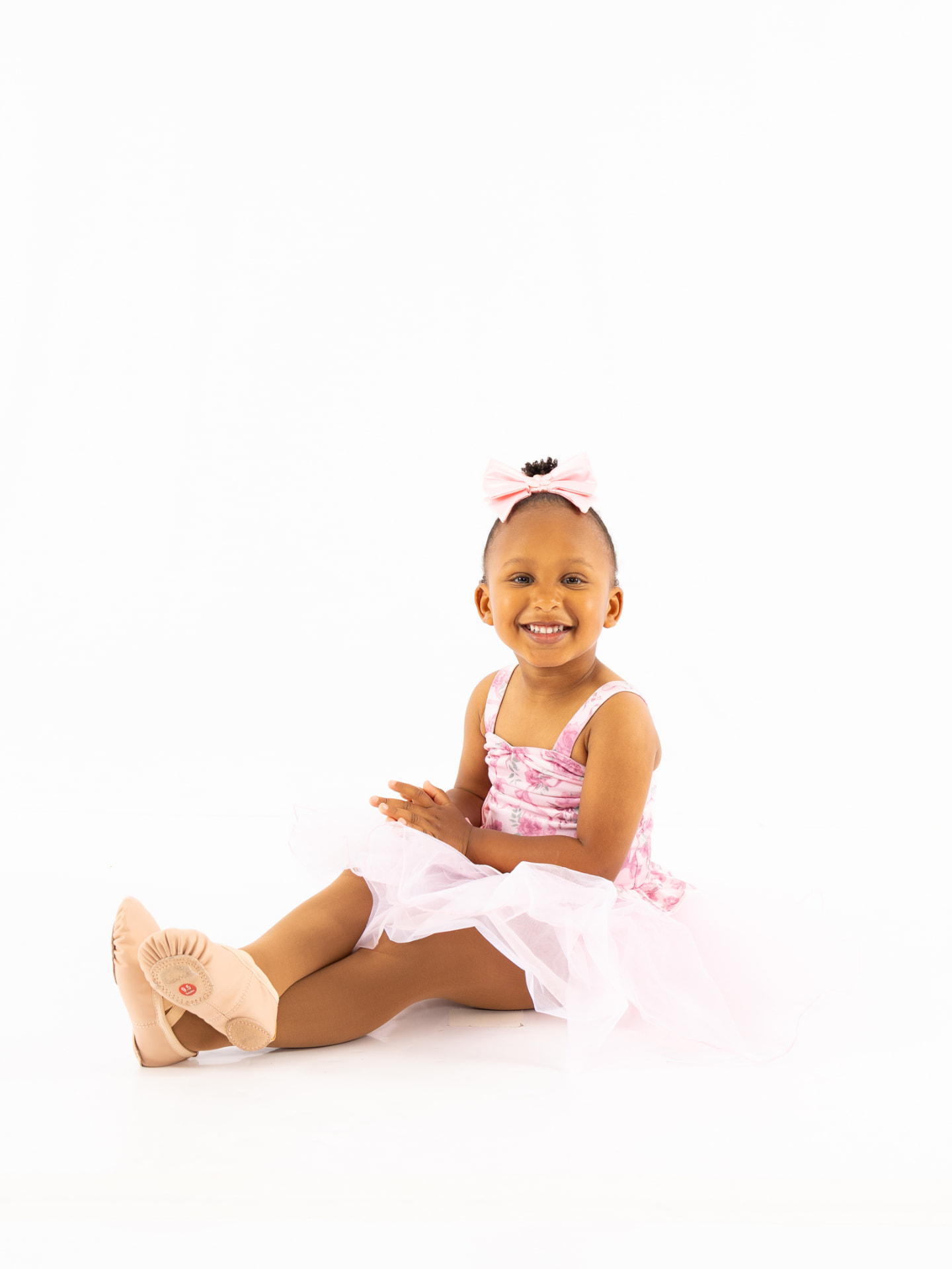 A smiling toddler dancer in a pink tutu sitting on the studio floor