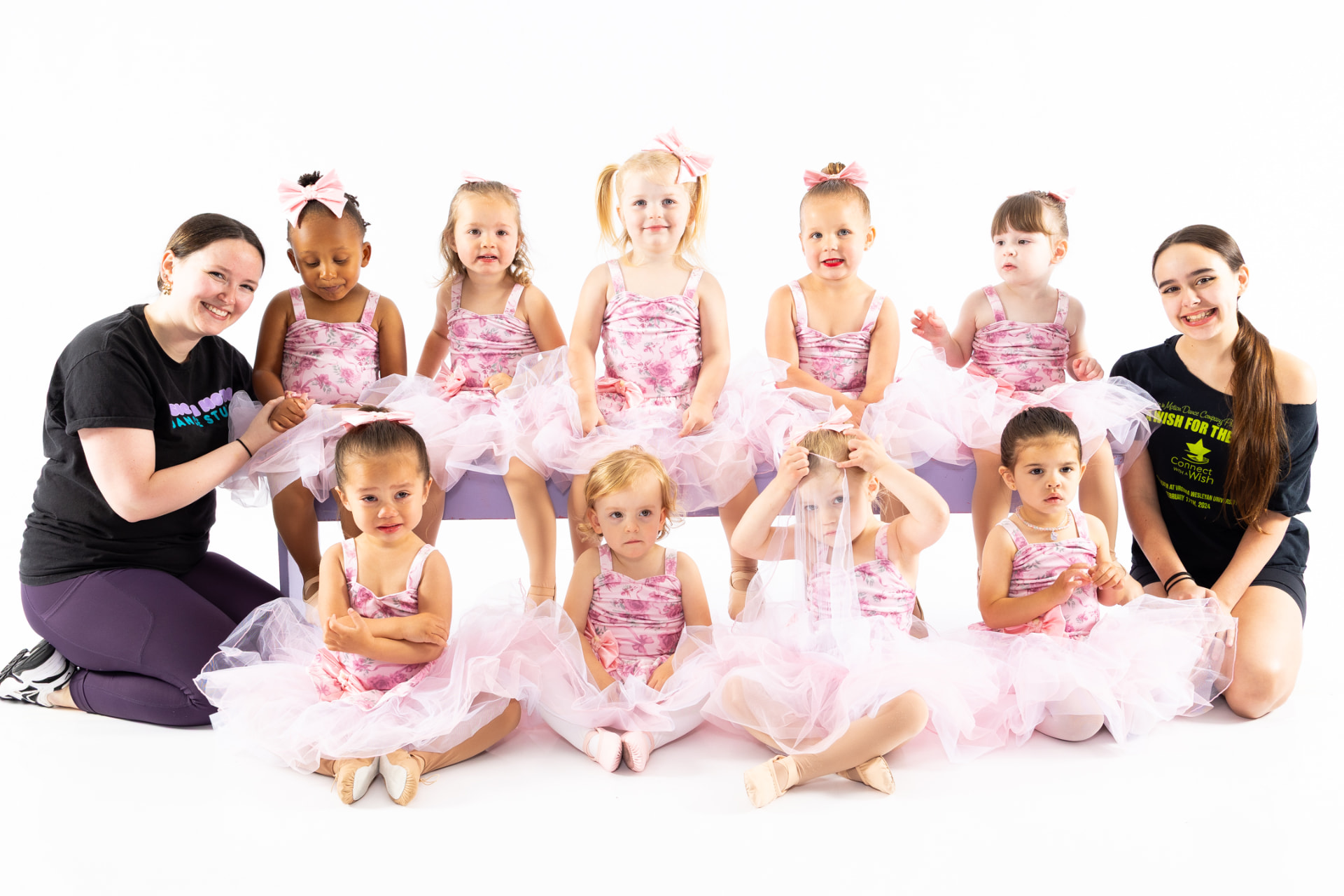 A preschool ballet class in pink costumes with two teachers on a lavender bench
